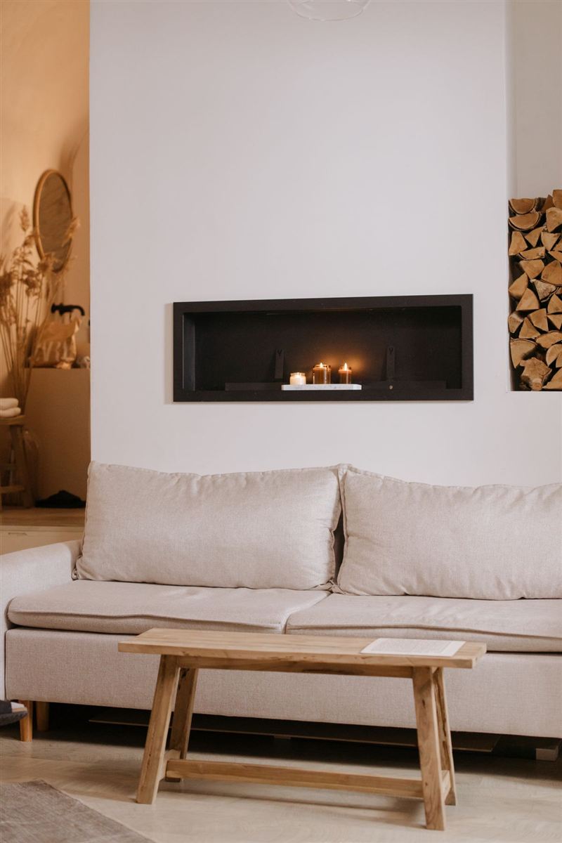 Cream sofa beneath a built-in fireplace niche lit with candles, a light wood bench used as a coffee table, and a stack of firewood, in warm ambient light.