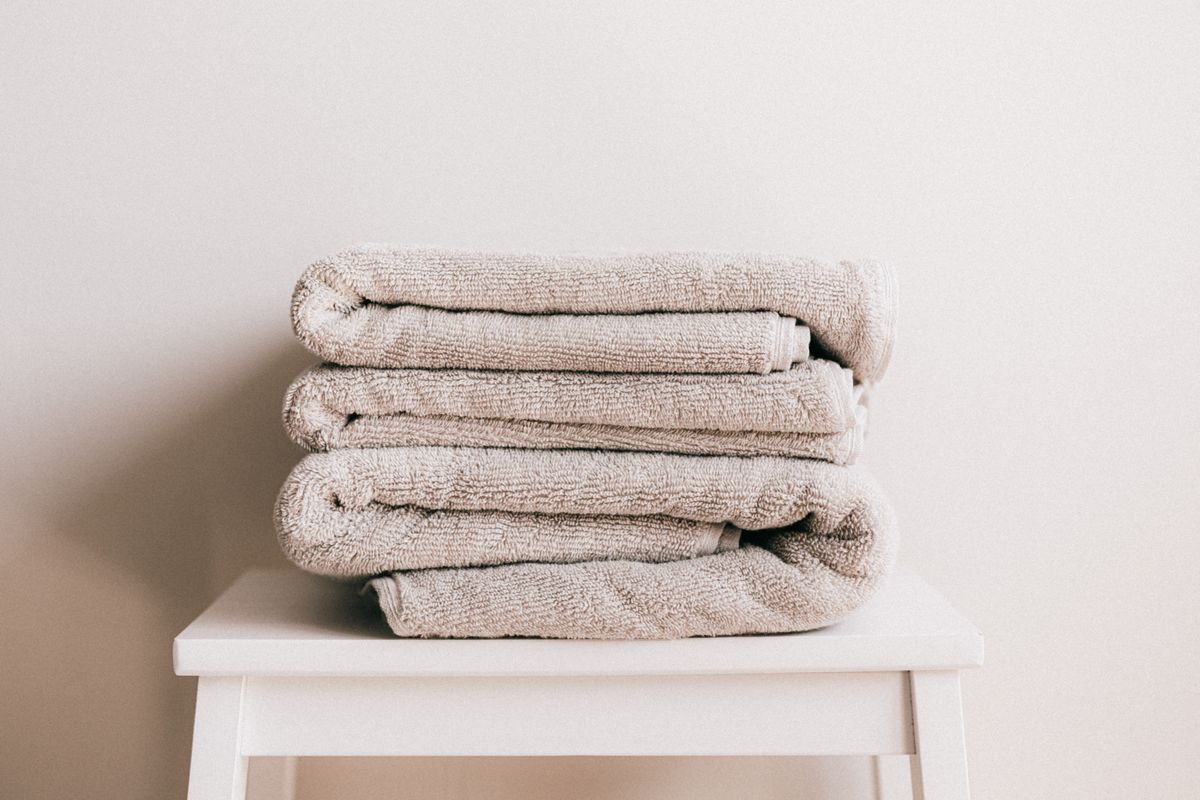 A neat stack of four rolled, oatmeal-beige towels on a white stool against a plain warm-white wall.