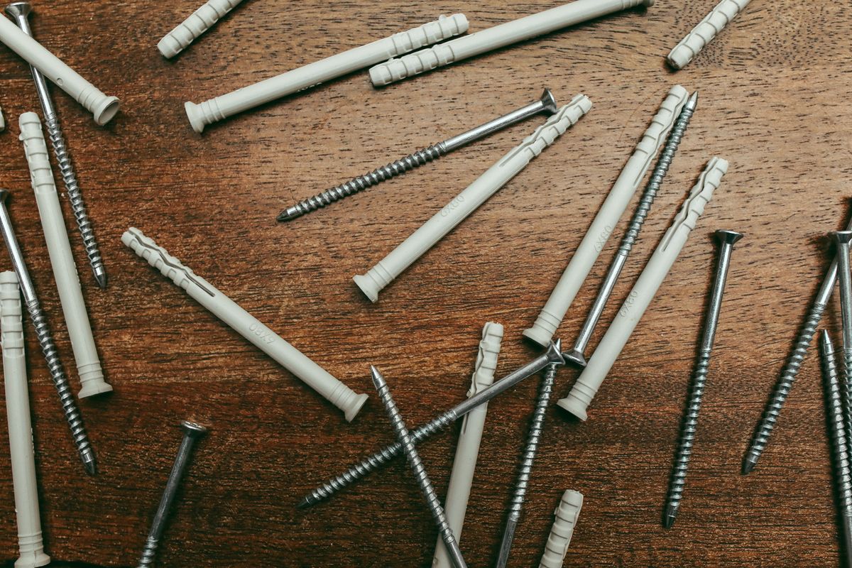 Overhead macro shot of metal screws and tan plastic wall anchors scattered across a warm wood surface.