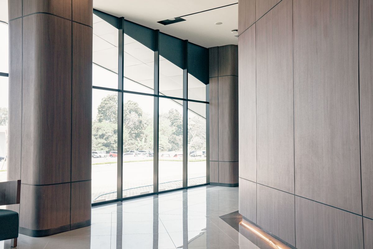 Bright modern commercial lobby with warm wood-panel walls, floor-to-ceiling glass, a polished reflective floor, and trees visible outside.