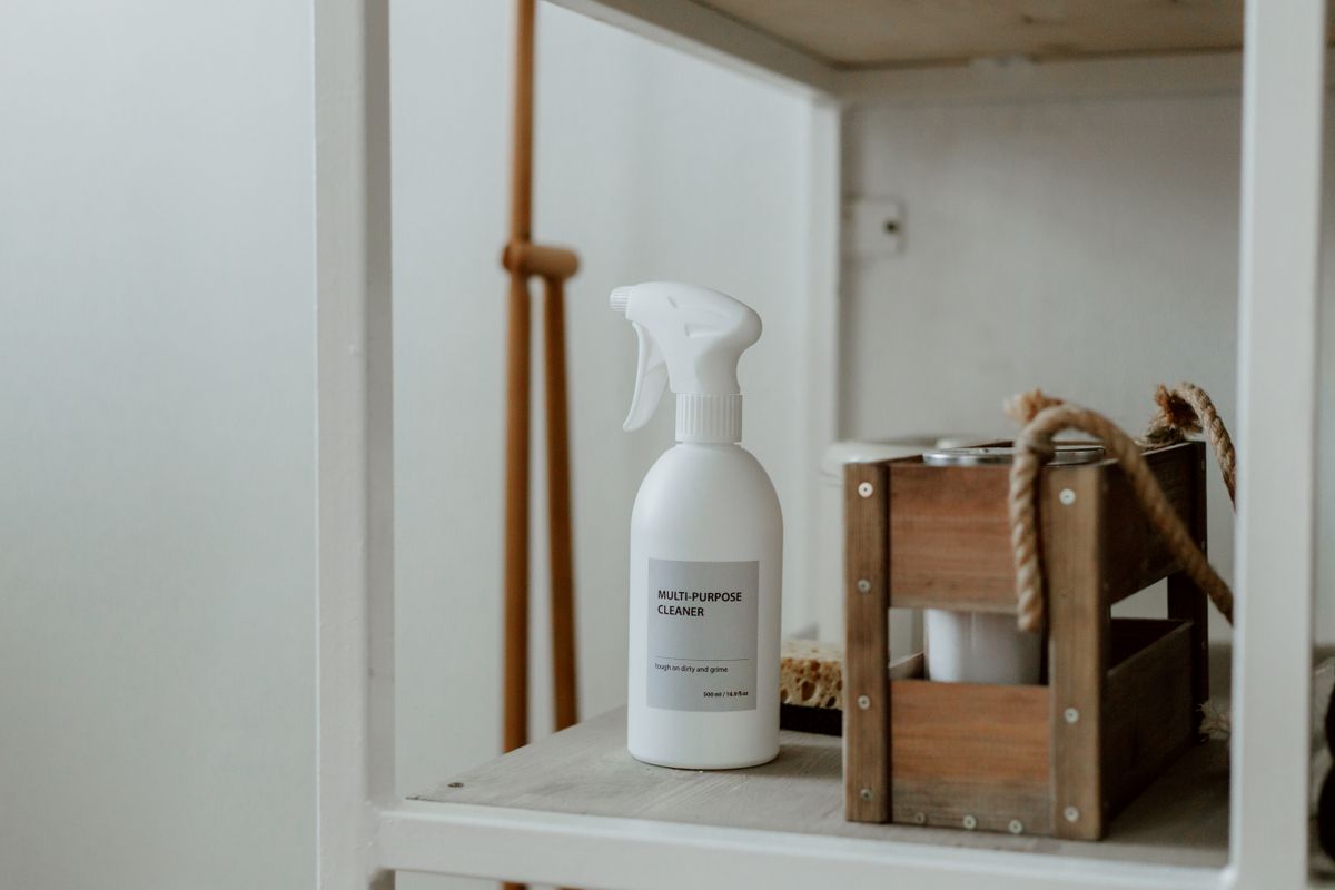 A plain white spray bottle labeled 'multi-purpose cleaner' on a white shelf next to a small wooden crate caddy with a rope handle.