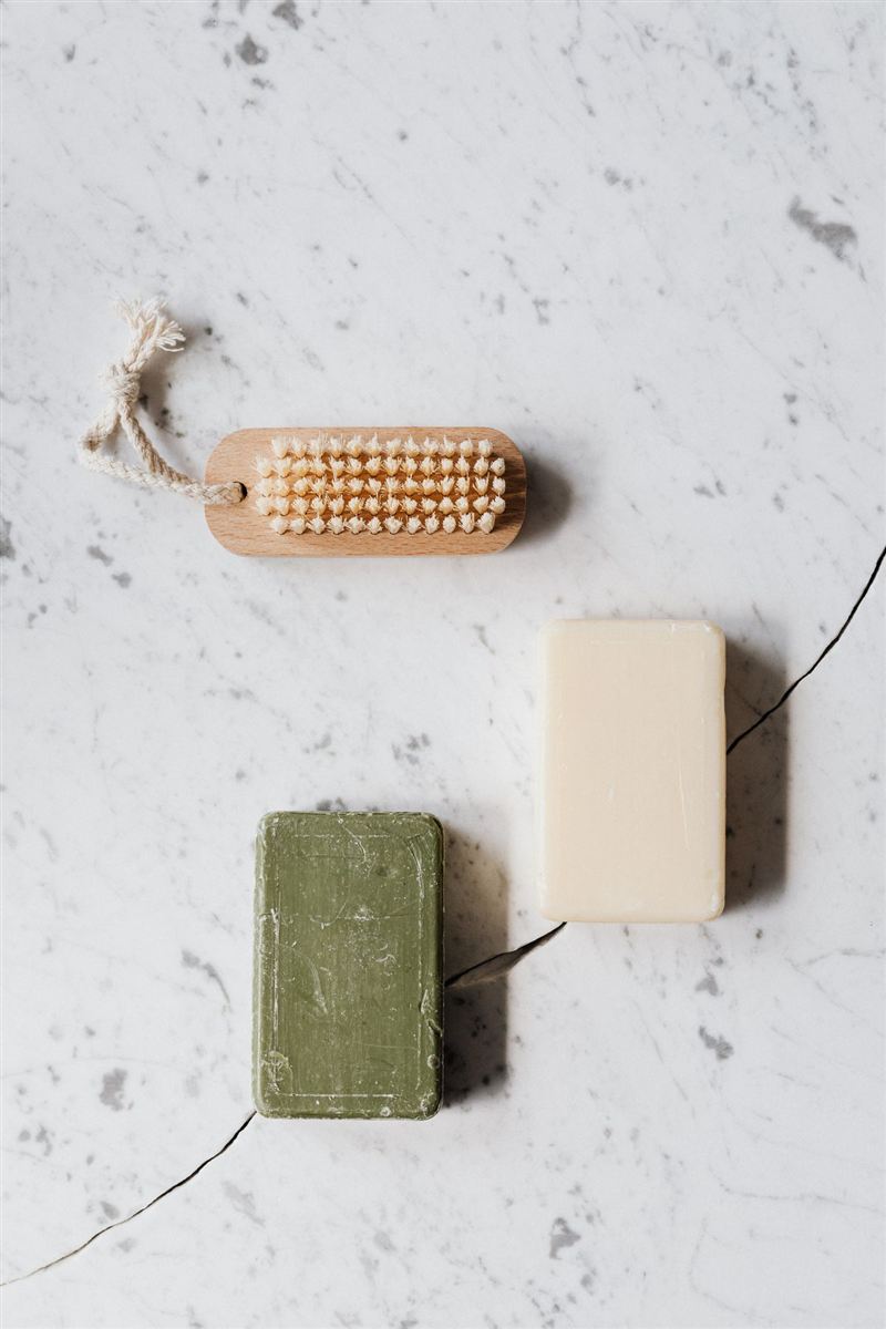 Flat lay on white marble of a wooden nail brush with cream bristles, a cream soap bar, and a small olive-green soap bar.