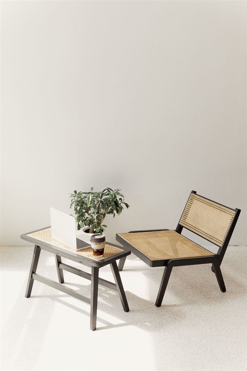 Styled minimalist workspace with a black-framed rattan chair and matching cane side table holding a laptop, a small potted plant, and a coffee cup, on a pale terrazzo floor.