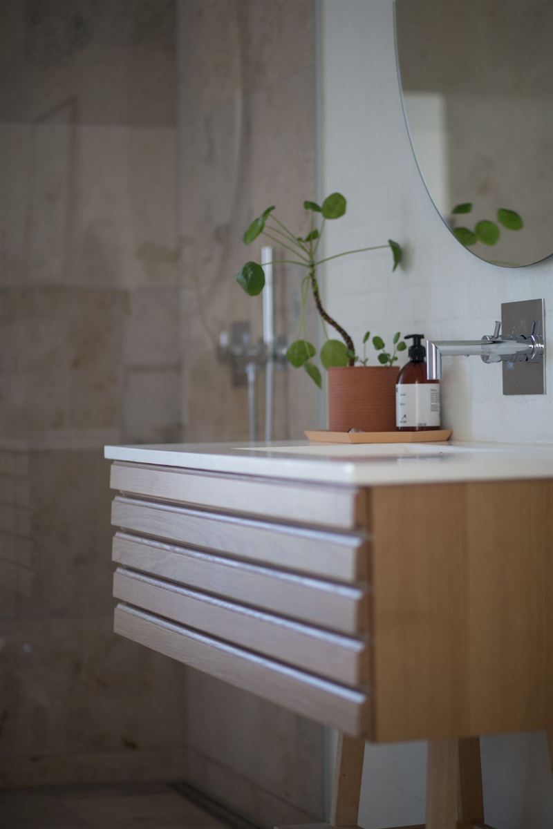 Spa-style bathroom detail: a fluted light-wood vanity, round mirror, wall-mounted chrome faucet, a small potted plant, and a bottle of soap, against a warm stone-textured wall.
