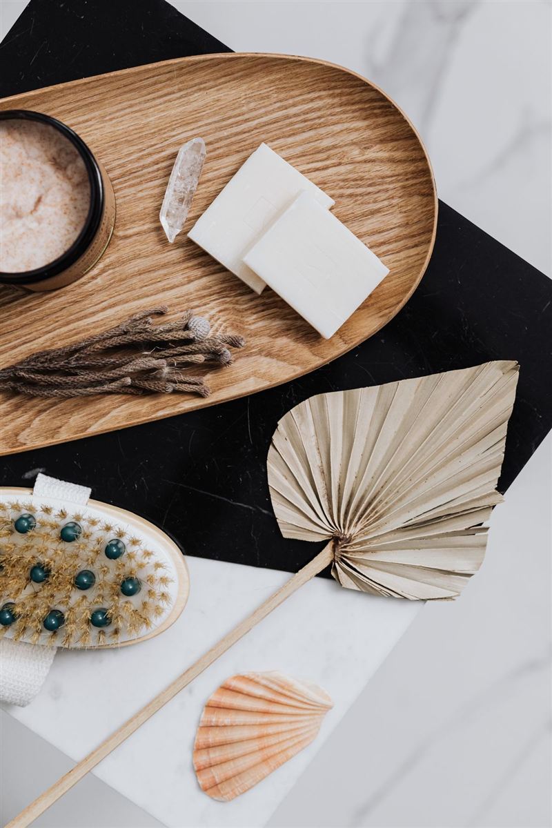 Styled flat lay on black and white marble: a round wood tray holding a jar of scrub, a crystal point, cream soap bars, and dried seed pods, beside a dried palm frond, a small shell, and a bristle brush.