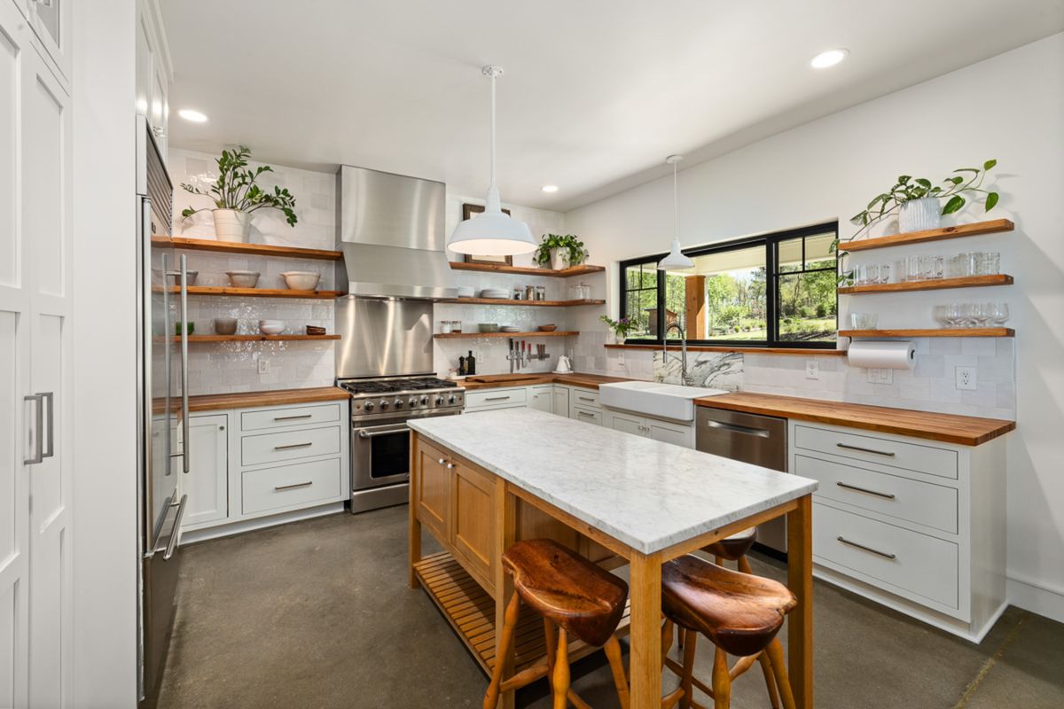 Bright kitchen with white cabinetry, open wood shelving holding dishware and potted plants, a marble-topped island with wood stools, and a window view of trees.