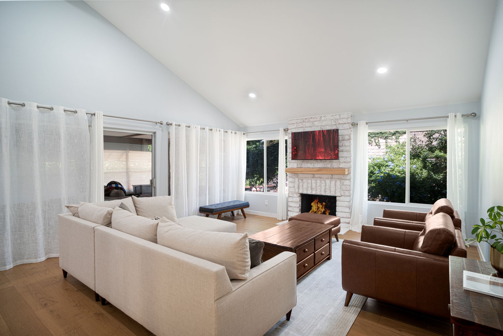 Sunlit living room with a cream sectional sofa, cognac leather armchairs, a wood coffee table, and a white brick fireplace; large windows show greenery outside.