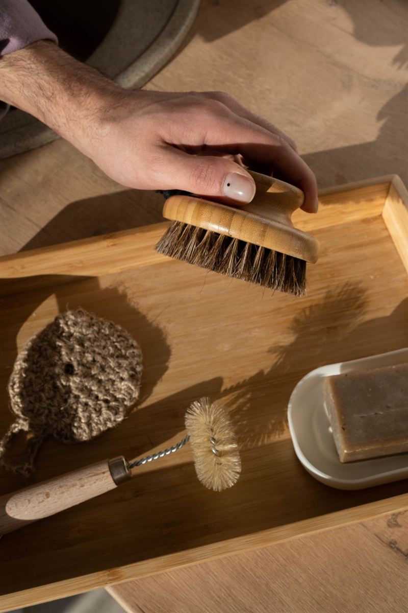 A hand picking up a wood-and-bristle bath brush from a bamboo tray that also holds a woven pad, a small wooden-handled brush, and a bar of soap on a dish.